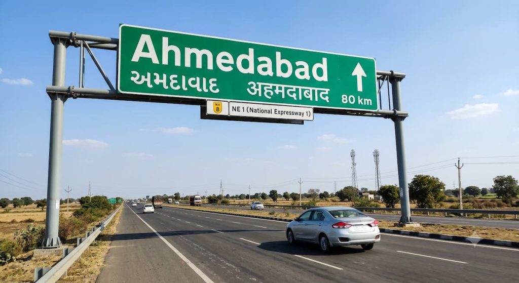 Best Connecting Airports for Rajkot to Dubai Flights 2 A photorealistic overhead road sign gantry on National Expressway 1 (NE 1) in India. The large green sign displays 'Ahmedabad' in large English, Gujarati (અમદાવાદ), and Hindi (अहमदाबाद) script, with an upward-pointing arrow and a distance of '80 km'. A smaller sign below indicates 'NE 1 (National Expressway 1)' with the Indian highway shield. The highway has multiple lanes with cars driving on it, running through a dry, open rural landscape under a clear blue sky.